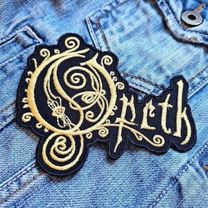May include: A black embroidered patch with the word "Opeth" in a stylized font. The patch features a gold-colored ornate design with swirls and a floral element. The patch is sewn onto a denim jacket.