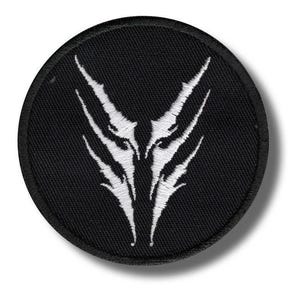 May include: A round, black embroidered patch featuring a white, stylized design. The design is symmetrical, with sharp, angular shapes radiating outwards from a central point. The patch has a black border and a textured appearance.