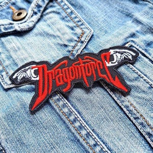 May include: An embroidered patch featuring the word "Dragonforce" in red, stylized lettering on a black background. The patch has white wing designs and is attached to a light blue denim jacket.