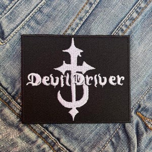 Puede incluir: Parche rectangular negro con las palabras "DevilDriver" en fuente gótica blanca, sobre un diseño estilizado de cruz y media luna. El parche está sobre un fondo de mezclilla.