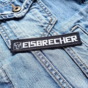 Op de afbeelding: Een rechthoekige zwart-wit geborduurde patch met het woord "EISBRECHER" in witte hoofdletters. De patch heeft een kleine afbeelding aan de linkerkant. Het is genaaid op een lichtblauwe spijkerjas.