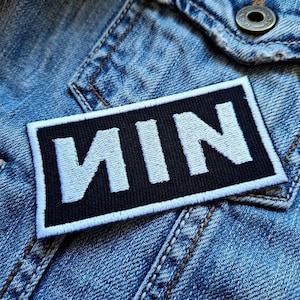 May include: A rectangular black patch with the letters "NIN" in white, embroidered on a denim jacket. The patch has a white border and is sewn onto the blue denim fabric. The jacket has a button and visible stitching.