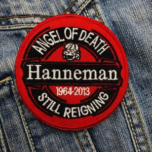 Könnte beinhalten: Ein runder, roter und schwarzer Aufnäher mit den weißen Worten "ANGEL OF DEATH" und "HANNEMAN". Der Aufnäher enthält auch den Text "1964-2013 STILL REIGNING" und eine Totenkopf-Grafik. Der Aufnäher befindet sich auf einem Jeansstoff-Hintergrund.