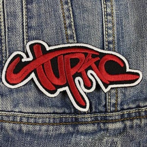 Puede incluir: Un parche bordado en rojo y negro con la palabra "UPRC" en una escritura estilizada, contorneada en blanco. El parche está cosido sobre una chaqueta vaquera azul, mostrando la textura de la tela y las costuras.