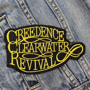 Könnte beinhalten: Ein schwarzer Aufnäher mit gelbem gesticktem Text, der "Creedence Clearwater Revival" lautet. Der Aufnäher hat eine dekorative Form und ist auf eine Jeansjacke genäht.