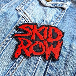 Könnte beinhalten: Ein schwarz-roter, gestickter Aufnäher mit den Worten "SKID ROW" in einer kräftigen, stilisierten Schriftart. Der Aufnäher ist auf eine hellblaue Jeansjacke genäht und zeigt das Design und die Textur des Aufnähers. Ein Rock'n'Roll-Accessoire.