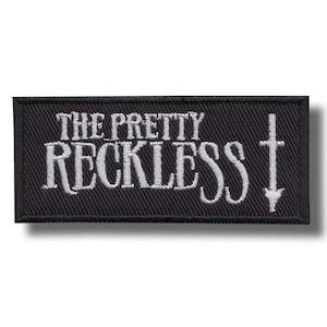 May include: Black rectangular patch with the text "THE PRETTY RECKLESS" in white, stylized lettering. A white cross with a downward arrow is on the right side. The patch has a woven texture and a white border.