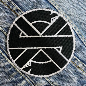 Könnte beinhalten: Ein runder, schwarz-weißer gestickter Aufnäher mit einem geometrischen Design. Der Aufnäher hat eine schwarze Mitte mit weißen Linien, die eine stilisierte Form bilden, und einen weißen Rand. Der Aufnäher ist auf eine Jeansjacke genäht.
