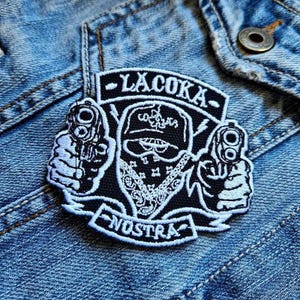 Könnte beinhalten: Schwarz-weißer gestickter Aufnäher mit dem Text "LACOKA NOSTRA". Der Aufnäher zeigt eine Figur mit Bandana, die zwei Pistolen hält. Der Aufnäher ist auf eine blaue Jeansjacke genäht.