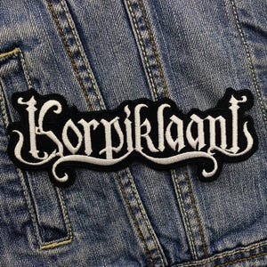 Könnte beinhalten: Ein schwarz-weiß gestickter Aufnäher mit dem Wort "Korpiklaani" in einer verzierten Schriftart. Der Aufnäher hat eine geschwungene Form und ist auf einer Jeansjacke platziert.