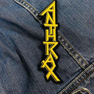 Può includere: Una toppa ricamata gialla e nera con la scritta "ANTHRAX" in un carattere stilizzato. La toppa è rettangolare ed è cucita su una giacca di jeans.