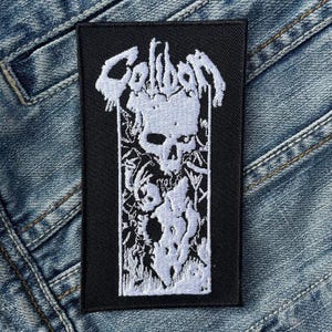 Puede incluir: Parche rectangular negro con un diseño de calavera blanca y la palabra "Caliban" en letras estilizadas. La calavera está rodeada de intrincados patrones blancos similares a flores. El parche está sobre un fondo de mezclilla.