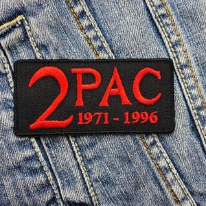 May include: A rectangular black patch with the red text "2PAC" and the dates "1971-1996" embroidered on it. The patch is placed on a light blue denim jacket.