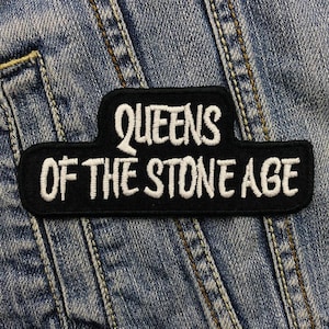 Könnte beinhalten: Ein schwarzer, gestickter Aufnäher mit dem weißen Schriftzug "QUEENS OF THE STONE AGE". Der Aufnäher hat eine rechteckige Form mit einer gebogenen Oberseite und liegt auf Jeansstoff.