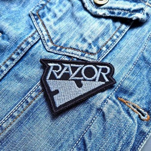 May include: A black and gray embroidered patch with the word "RAZOR" and a razor blade design. The patch is sewn onto a light blue denim jacket, with visible stitching and a button.