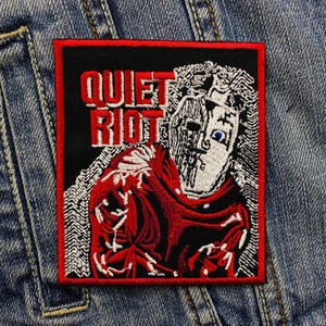 May include: Embroidered patch with a black background and a red border. The patch features the text "QUIET RIOT" in red block letters. A stylized illustration of a figure with a red cloak and a white face is also present.