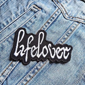 May include: A black embroidered patch with the word "lifelover" in white cursive script. The patch is sewn onto a light blue denim jacket, showcasing a casual, everyday style.