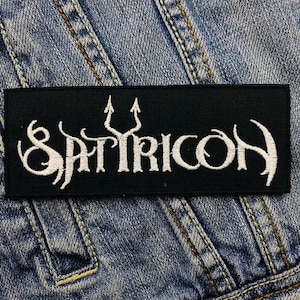 May include: A black rectangular patch with the word "SATYRICON" in white, stylized lettering. The patch features a trident symbol above the "I" in the word. The patch is sewn onto a denim jacket.