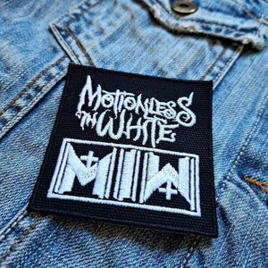 Op de afbeelding: Zwart vierkantige patch met witte geborduurde tekst "Motionless in White" boven "MIW" met kruissymbolen. De patch zit op een spijkerjasje.