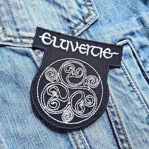 Op de afbeelding: Zwarte patch met witte geborduurde tekst "Eluveitie" boven een cirkelvormig ontwerp van drie wervelende vormen. De patch is op een spijkerjas genaaid.