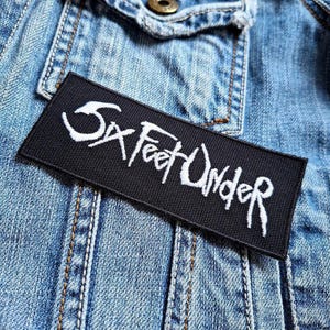 Puede incluir: Un parche rectangular negro con las palabras "Six Feet Under" bordadas en blanco, exhibido sobre una chaqueta vaquera. El texto tiene una fuente estilizada, escrita a mano. El parche es rectangular y tiene una textura tejida.