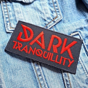 Könnte beinhalten: Ein rechteckiger schwarzer Aufnäher mit den Worten "DARK TRANQUILLITY" in Rot gestickt. Der Aufnäher ist auf eine hellblaue Jeansjacke genäht, mit sichtbaren Nähten und einer Tasche im Hintergrund.