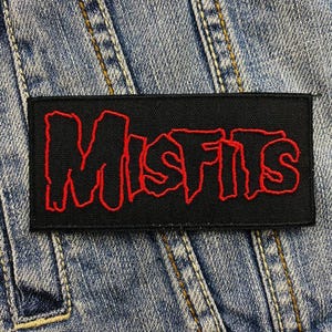 Puede incluir: Un parche rectangular negro con la palabra "Misfits" bordada en rojo con letras estilizadas. El parche está sobre un fondo de mezclilla azul claro.
