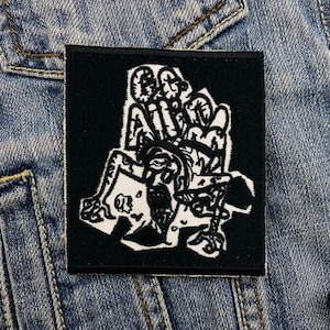 Puede incluir: Parche cuadrado negro con una ilustración gráfica blanca de una figura sentada en un trono. El parche está sobre un fondo de mezclilla azul.