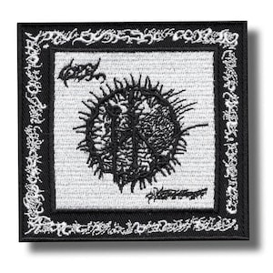 Puede incluir: Parche bordado cuadrado en blanco y negro. El parche presenta un diseño central negro y puntiagudo sobre un fondo blanco, con texto negro arriba y abajo. El borde es negro con un patrón decorativo blanco.