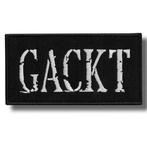 May include: Black rectangular patch with the word "GACKT" embroidered in white, distressed-style lettering. The patch has a black border and is likely designed to be sewn or ironed onto clothing or accessories.