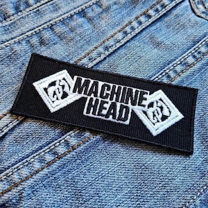 Op de afbeelding: Zwart rechthoekig embleem met de woorden "MACHINE HEAD" in witte, gestileerde tekst. Het embleem heeft witte ruitvormige ontwerpen aan elk uiteinde. Het embleem is op een blauw spijkerjasje genaaid.