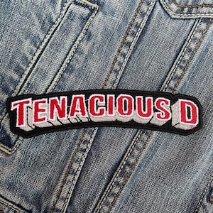 Peut inclure: Un écusson brodé noir, rouge et blanc avec le texte "TENACIOUS D" en relief. L'écusson rectangulaire est cousu sur une veste en jean bleu clair.