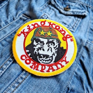 May include: Embroidered patch with a yellow border and the words "King Kong Company" in red. The patch features a gorilla wearing a military-style helmet with two gold stars. The patch is sewn onto a denim jacket.