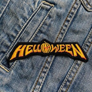 Może przedstawiać: Czarna i żółta haftowana naszywka z napisem "HELLOWEEN" i wzorem dyni pośrodku. Naszywka jest przyszyta do niebieskiej dżinsowej kurtki, ukazując szczegóły haftu i fakturę materiału.