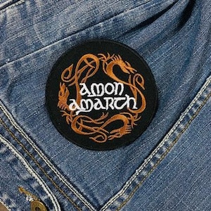 May include: A round, black patch with the words "amon amarth" in white and orange, surrounded by an orange dragon design. The patch is sewn onto a blue denim jacket.