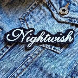 Peut inclure: Un écusson brodé noir et blanc avec le mot "Nightwish" dans une police stylisée. L'écusson est posé sur une veste en jean bleue, avec le texte en blanc et un bord noir.