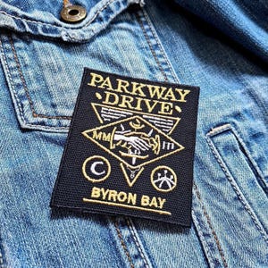 Op de afbeelding: Een zwart embleem met gouden letters en ontwerp, met de tekst "PARKWAY DRIVE" en "BYRON BAY". De patch is op een spijkerjas genaaid. Het ontwerp bevat een handdruk, een halve maan en andere symbolen.