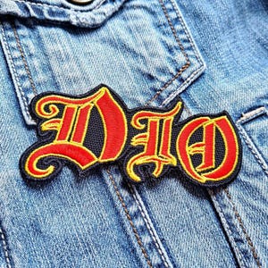 May include: An embroidered patch featuring the word "DIO" in red, with a yellow outline, on a black background. The patch is in a gothic font and is set against a denim jacket.