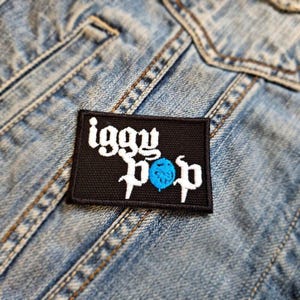 May include: A black rectangular patch featuring the name "Iggy Pop" in white, decorative lettering. A blue sphere is between the words "Pop". The patch is attached to a light blue denim jacket.