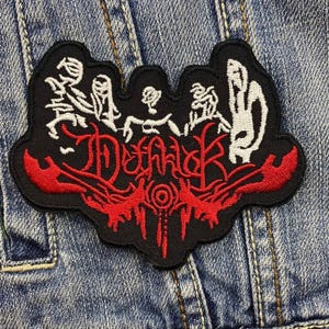 Puede incluir: Parche bordado negro con detalles rojos y blancos. El parche presenta texto estilizado y figuras abstractas. El parche está cosido a una chaqueta vaquera. El texto en el parche dice "Deafh".