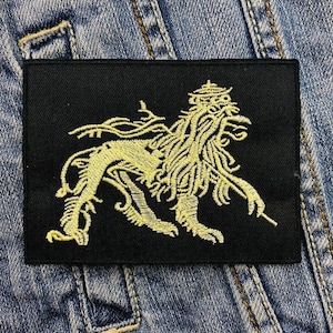 May include: Black rectangular patch featuring a gold embroidered lion design. The lion is depicted in a detailed, stylized manner with flowing mane and intricate details. The patch is likely for clothing or accessories.