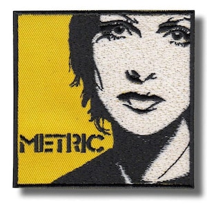 Pode incluir: Patch bordado com um retrato estilizado em preto e branco sobre um fundo amarelo. A palavra "METRIC" está escrita em letras pretas na seção amarela.
