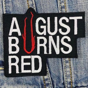 May include: Embroidered black patch featuring the text "AUGUST BURNS RED" in white, with a red flame graphic. The patch has a distinctive shape and is displayed on a denim fabric background.