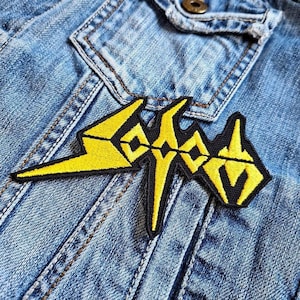 May include: A black and yellow embroidered patch with the word "SODOM" in a stylized font is sewn onto a light blue denim jacket. The patch features a bold, geometric design with yellow lightning bolt shapes and black outlines. The jacket has visible seams and a button.