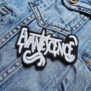 May include: A black and white embroidered patch with the word "EVANESCENCE" in a stylized font. The patch is rectangular with rounded edges and is sewn onto a denim jacket.