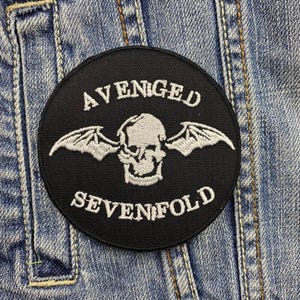 以下が含まれることがあります： 黒い円形のパッチで、白い刺繍の文字と翼のあるドクロのデザインが施されています。「AVENGED」と「SEVENFOLD」の文字が刺繍されています。パッチはデニムジャケットに縫い付けられています。