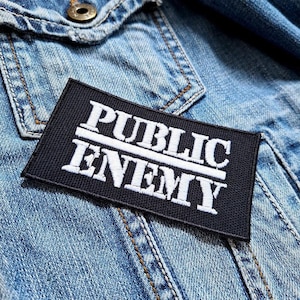 May include: A black rectangular patch with the words "PUBLIC ENEMY" embroidered in white. The patch is placed on a denim jacket, with the stitching and texture of the jacket visible.