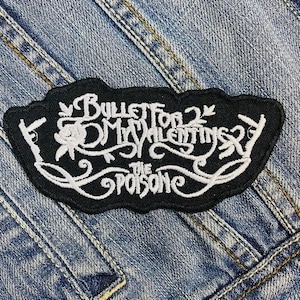 Könnte beinhalten: Schwarzer, gestickter Aufnäher mit dem Text "Bullet for My Valentine" in weißer Schrift. Der Aufnäher hat ein dekoratives Blumendesign und ist vor einem Jeanshintergrund platziert.