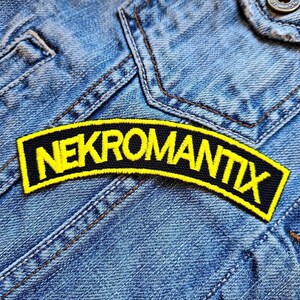 Pode incluir: Um patch bordado preto e amarelo com a palavra "NEKROMANTIX" em letras amarelas. O patch é arqueado e colocado sobre uma jaqueta jeans azul.