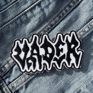 May include: An embroidered patch featuring the word "VADER" in black, outlined in white. The patch is on a blue denim background, with visible seams and stitching.